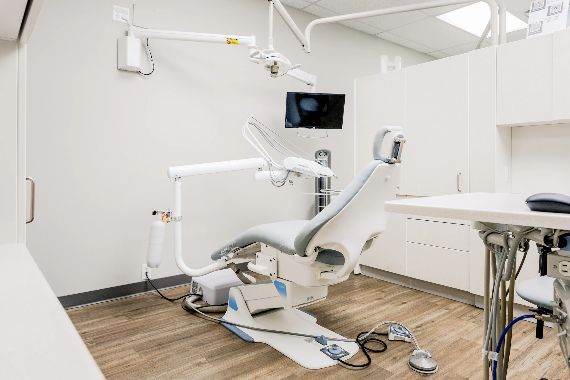 A modern dental chair beside a wall-mounted screen in a clean treatment room