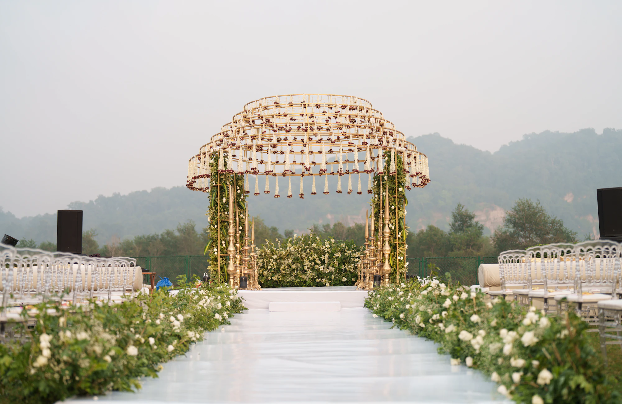 An outdoor wedding aisle lined with white flowers leading to a floral canopy, hills behind