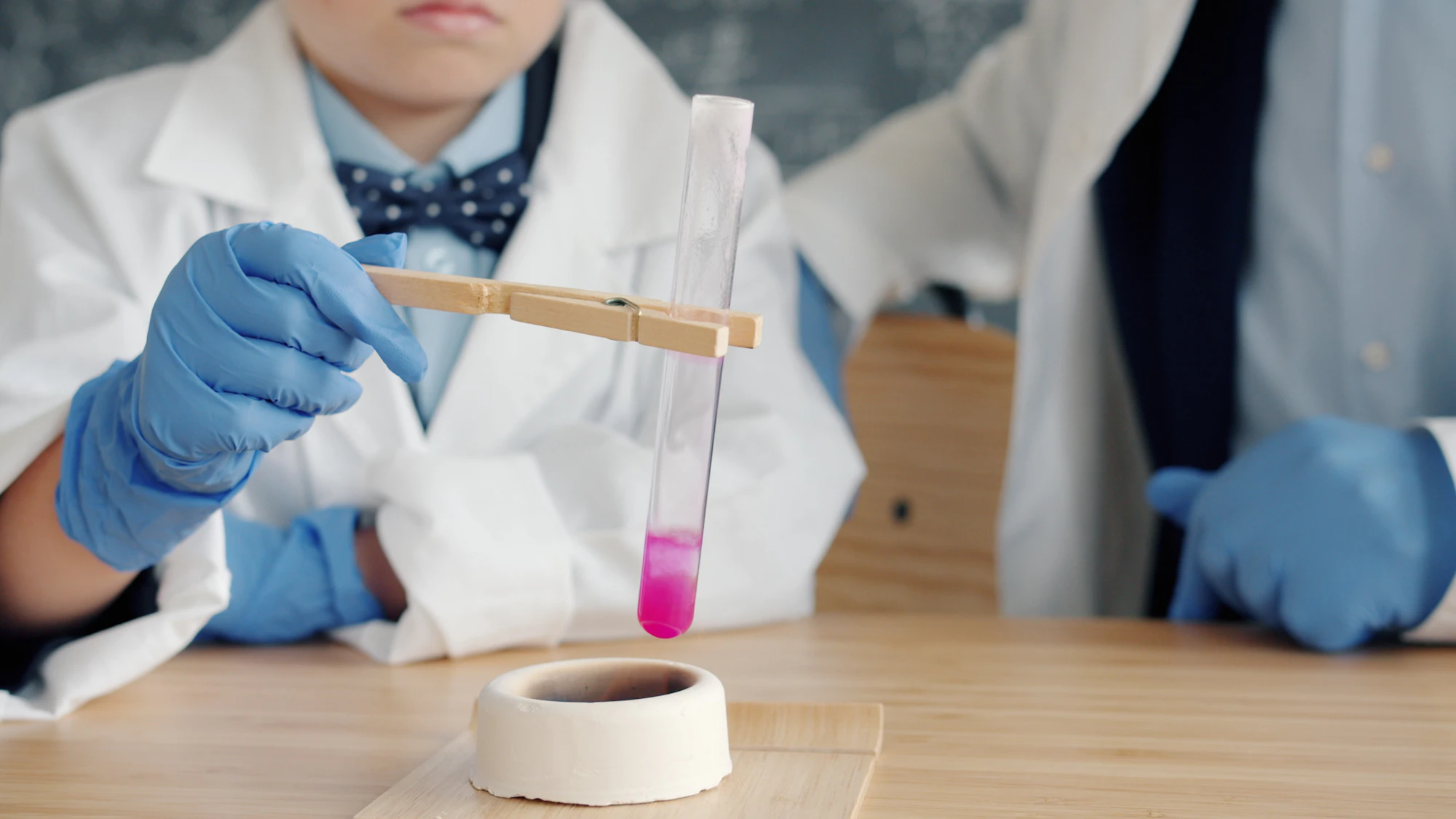 A child in a lab coat and gloves holding a test tube of pink liquid