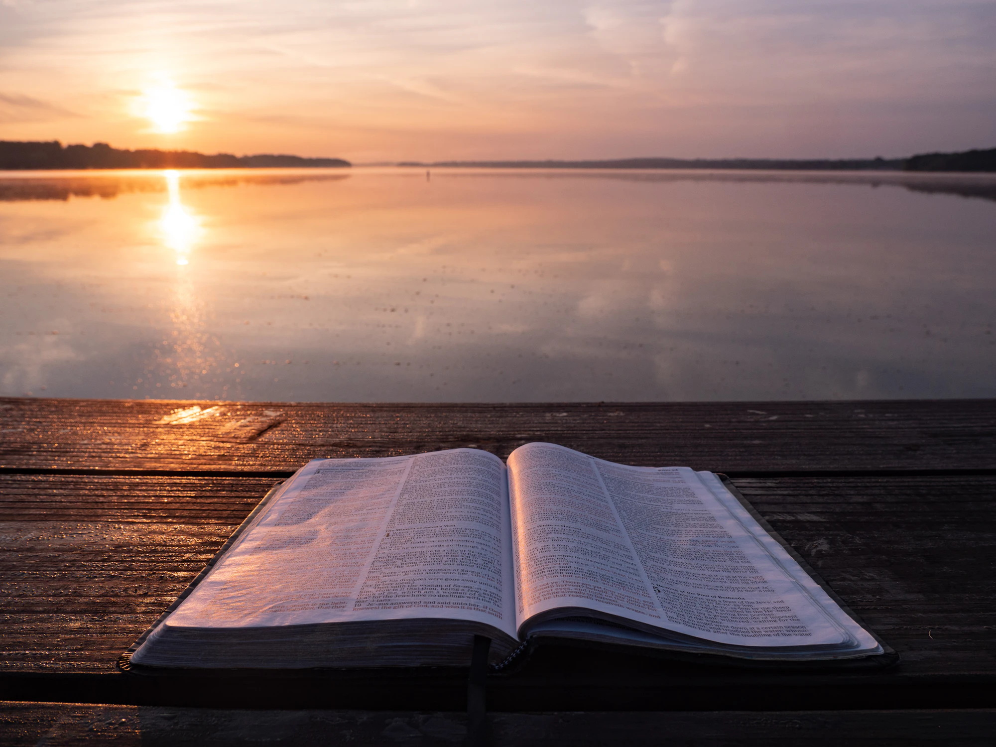 An open book on a wooden table by a lake at sunrise