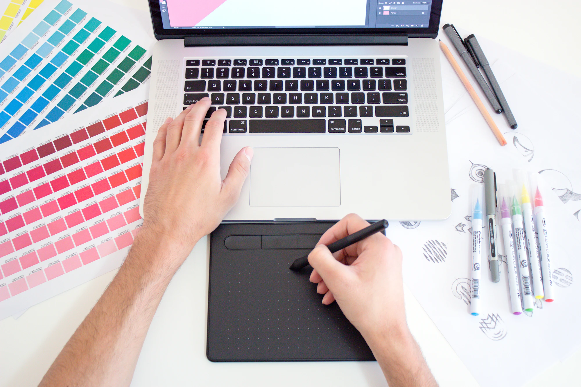 Hands using a drawing tablet beside a laptop and colour swatches